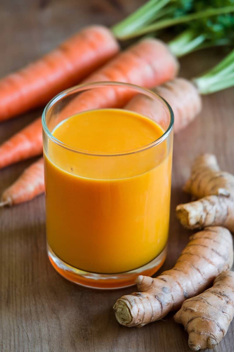 A glass of bright orange carrot and turmeric juice with whole carrots and turmeric roots in the background