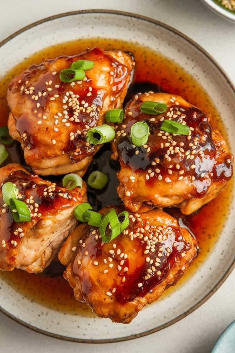 A plate of skinless boneless chicken thighs glazed with spicy honey soy sauce, garnished with sesame seeds and green onions.