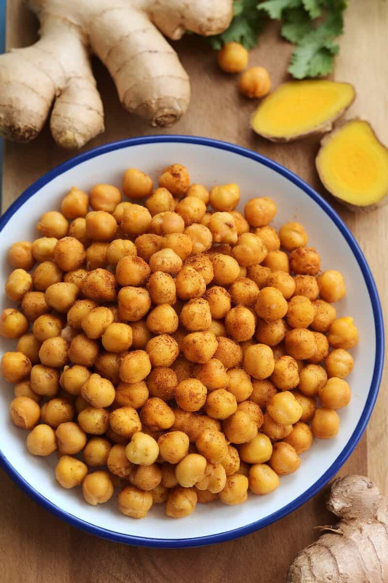 A bowl of roasted chickpeas with turmeric and ginger, surrounded by fresh ginger root and turmeric slices.