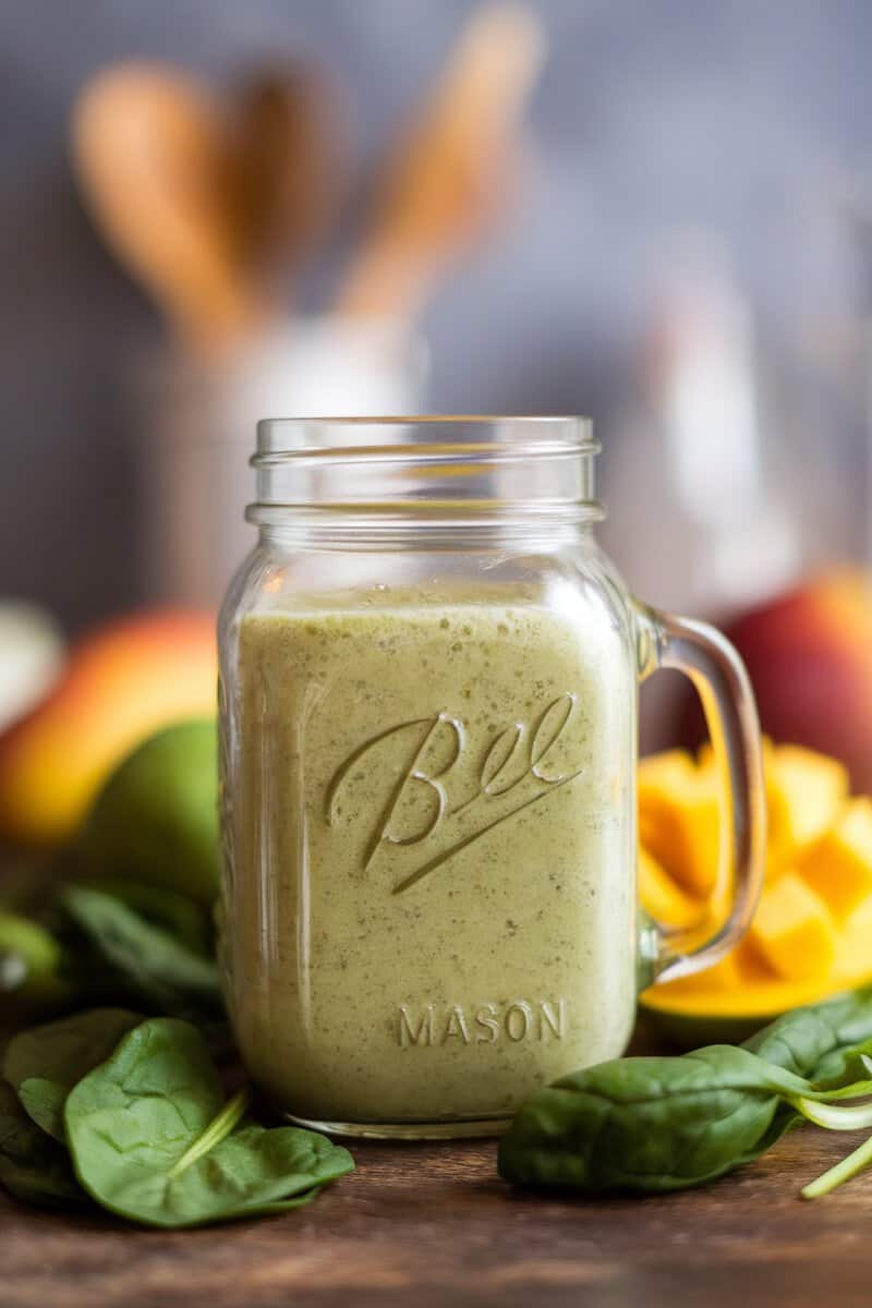 A refreshing mango spinach green smoothie in a glass jar, surrounded by fresh fruits and spinach leaves.